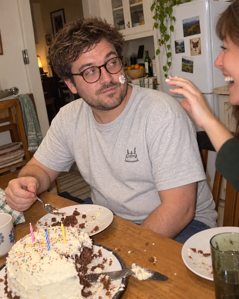 Studio concept: a birthday guest with icing on his cheek and a tiny cake embroidered on his grey tee