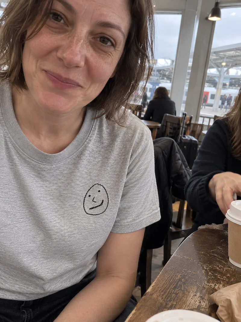 Studio concept: a very close casual phone portrait in a train-station café, wearing the small crooked face