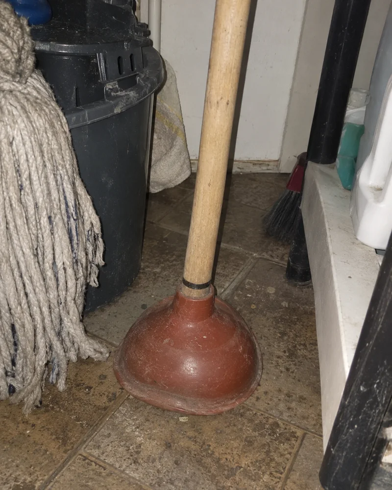 Studio concept: a worn brick-red rubber plunger in a cluttered cleaning cupboard