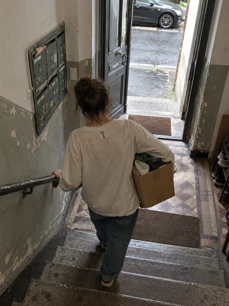 Studio concept: a person seen from behind carrying a moving box down a worn apartment stairwell