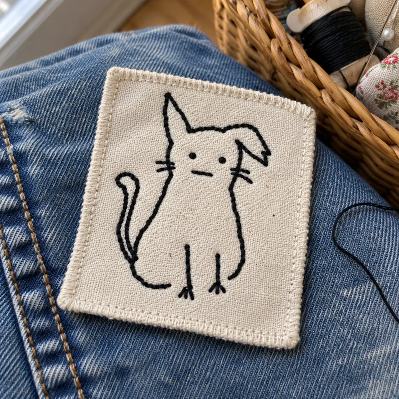 Studio concept: a small crooked-ear cat patch on worn denim beside a sewing basket