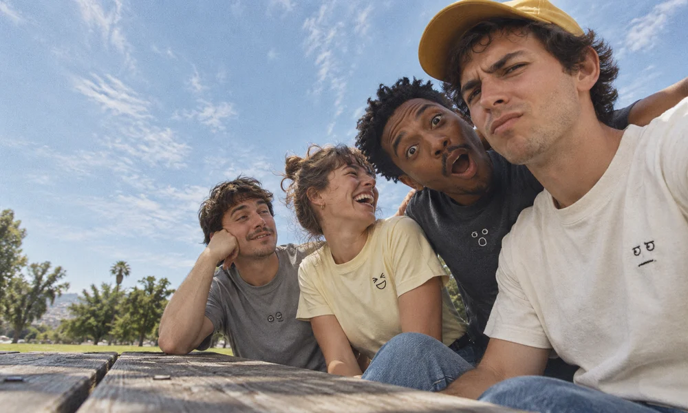 Studio concept: four friends sharing a joke at a picnic table, with small embroidered expressions on their tees